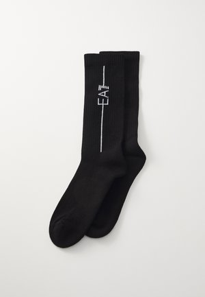 Chaussettes noires à côtes avec un logo blanc et un simple détail de ligne courant verticalement sur le côté. Texture lisse, design à la cheville.