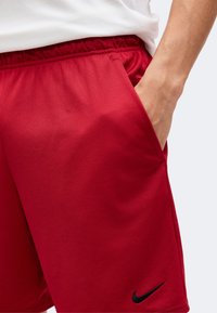 Personne portant un short rouge Nike avec la main dans la poche, taille élastique visible et une partie d'une chemise blanche visible au-dessus du short.