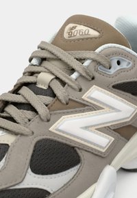 Harmaa ja beige lenkkari, jossa on teksturoitu verkkomateriaali, kumipohja ja näkyvä valkoinen 'N' logo. Ominaisuuksiin kuuluu monimutkainen nauhakuosisuunnittelu ja pehmustettu kaulus.
