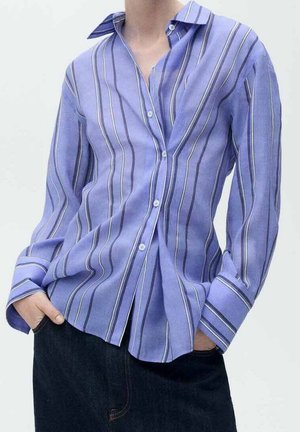 Henkilö, jolla on päällään vaaleanlila napitettava paita, jossa on pystysuorat mustavalkoiset raidat, kädet taskuissa, yhdistettynä tummiin housuihin.