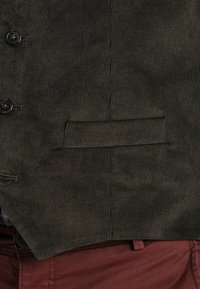 Gilet in corduroy verde scuro con una superficie texturizzata, dotato di una tasca sul petto e due bottoni scuri. Abbinato a pantaloni bordeaux.