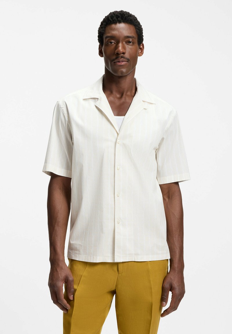 Homme portant une chemise crème à manches courtes boutonnée sur un sous-vêtement blanc et un pantalon jaune moutarde, debout devant un fond blanc uni.