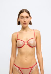 Διαφανές γυμνό σουτιέν με κόκκινες λεπτομέρειες και λεπτές λωρίδες, με κεντρική διακοσμητική λεπτομέρεια. Συμπληρωματικό διαφανές γυμνό εσώρουχο.