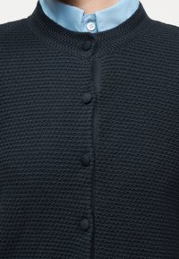 Κοντινό πλάνο ενός ατόμου που φοράει υφασμάτινο σκούρο μπλε ζακέτα με κουμπιά καλυμμένα με ύφασμα πάνω από ένα ανοιχτό μπλε πουκάμισο με γιακά.