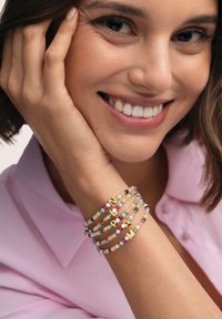 Ensemble de bracelets à perles dans des couleurs pastel avec des lettres accentuées d'or, présentant diverses textures et de petites perles rondes sur un fond clair.