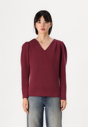 Top bordeaux a maniche lunghe con scollatura a V, caratterizzato da spalle gonfie e una texture liscia, abbinato a jeans di denim blu chiaro.