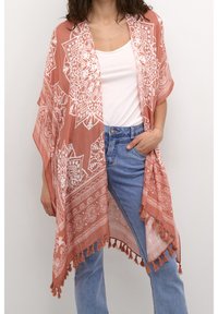 Lichtgewicht koraalkleurige sjaal met witte mandalapatronen; heeft franjes langs de randen. Draagt over een wit shirt en blauwe spijkerbroek.