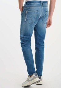 Blauwe denim jeans met een slim fit, voorzien van twee achterzakken en subtiele stiksel details. Gecombineerd met lichtgrijze sportschoenen.