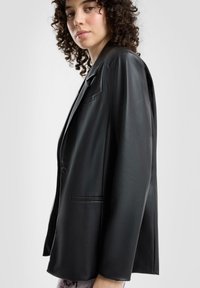 Czarna marynarka z ekologicznej skóry o dopasowanym kroju, z zapięciem na jeden guzik i usztywnionym ramionami. Posiada boczną kieszeń oraz gładką fakturę.