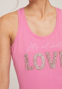 Vaaleanpunainen hihaton toppi, joka on valmistettu pehmeästä puuvillasta. Edessä tekstit "Juhlitaan" valkoisella käsinkirjoitetulla fontilla ja "RAKASTA" suurilla, hohtavilla hopeakirjaimilla.