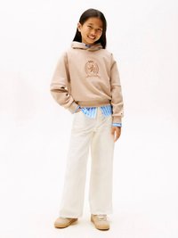 Ragazzina sorridente, indossa una felpa beige con uno stemma, una maglietta a righe blu sotto, pantaloni bianchi larghi e sneakers beige, in piedi con una mano in tasca.