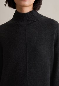 Temno siva volnena pulover s turtleneck ovratnikom in mehko, teksturirano površino. Ima srednji šiv, ki teče navpično po sprednji strani.