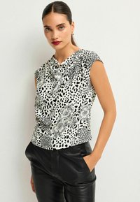 Top a maniche corte bianco con motivi floreali e maculati neri. Tessuto morbido con scollatura drappeggiata, abbinato a pantaloni in pelle nera.