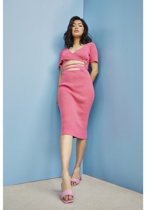 Abito midi rosa aderente a coste con dettagli cut-out e maniche corte, abbinato a tacchi aperti rosa coordinati su uno sfondo blu.