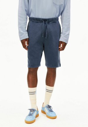 Man draagt marineblauwe shorts met trekkoord, lichtblauw overhemd met lange mouwen, crèmekleurige sokken met strepen en lichtblauwe sneakers met rubberen zolen.