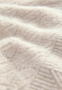 Tessuto lavorato a maglia beige chiaro che mostra un motivo ondulato e testurizzato con una consistenza morbida e soffice. Presenta dettagli a costine lungo il bordo.