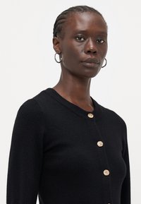 Cardigan negru tricotat cu guler rotund și butoane decorative aurii. Prezintă mâneci lungi ajustate și o textură moale.