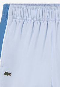 Ανοιχτό μπλε μαγιό σορτς με αντίθετη μπλε πλευρική λεπτομέρεια, ελαστική μέση και πράσινο κεντημένο λογότυπο κροκοδείλου στο αριστερό πόδι.