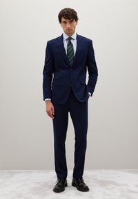 Junge Mann steht in einem marineblauen Anzug mit einem hellblauen Hemd, gestreifter grüner Krawatte und schwarzen Business-Schuhen vor einem schlichten hellen Hintergrund.