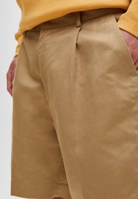 Los pantalones cortos de algodón beige cuentan con un corte relajado, con pliegues delanteros y bolsillos, y están detallados con una textura suave y costuras limpias.