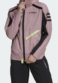 Ljusrosa vattentät jacka med svarta detaljer, dragkedjefickor och justerbar huva. Har en pastellgul dragkedja och logotyp. Perfekt för utomhusaktiviteter.