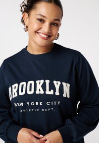 Sweatshirt bleu marine avec "BROOKLYN NEW YORK CITY ATHLETIC DEPT." en lettres blanches brodées. Présente un col et des poignets côtelés.