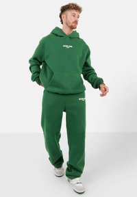 Man met krullend haar, gekleed in een groene hoodie en bijpassende joggingbroek, met witte sneakers, staande en zijwaarts kijkend.