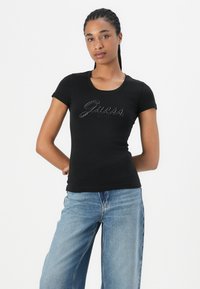 Black fitted t-shirt with short sleeves, featuring a decorative "Guess" logo in rhinestones. Paired with light blue denim wide-leg jeans.