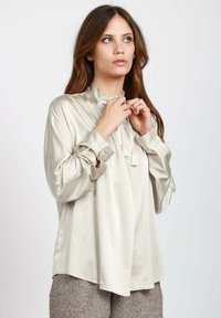 Blusa beige simile alla seta con collo alto, maniche lunghe e polsini abbottonati; presenta frange decorative al collo e una vestibilità ampia.