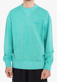 Homme portant un sweat-shirt turquoise avec un petit logo "element" sur la poitrine, associé à un pantalon noir, debout devant un fond blanc.