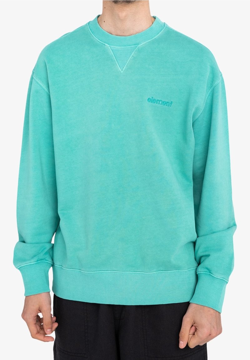 Homme portant un sweat-shirt turquoise avec un petit logo "element" sur la poitrine, associé à un pantalon noir, debout devant un fond blanc.