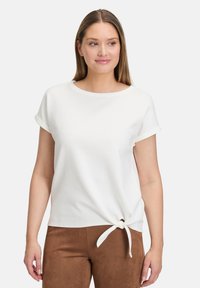 Biała bluzka z krótkim rękawem z podwiniętymi mankietami, z węzłem z przodu. Materiał ma subtelną teksturalną strukturę.