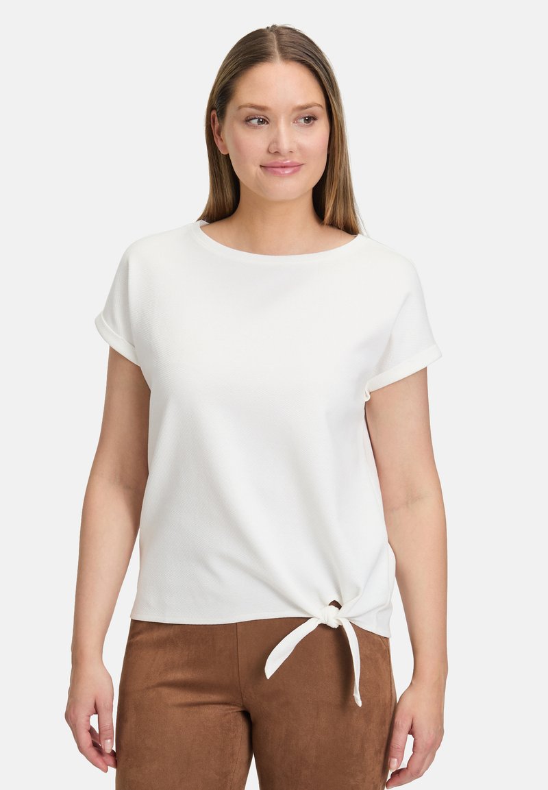 Biała bluzka z krótkim rękawem z podwiniętymi mankietami, z węzłem z przodu. Materiał ma subtelną teksturalną strukturę.
