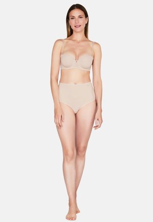 Donna in piedi a piedi nudi che indossa un reggiseno beige senza spalline e slip a vita alta senza cuciture su sfondo bianco.