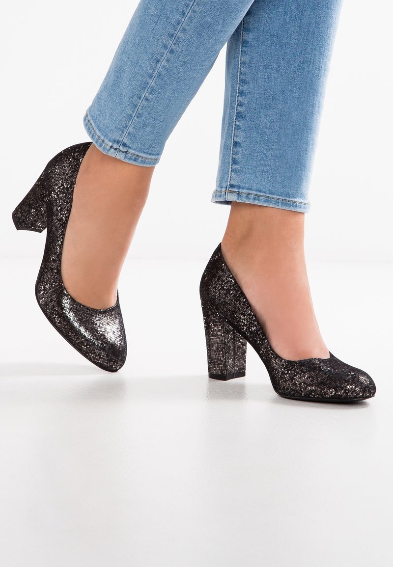 Pieds portant des chaussures à talons blocs brillantes à motif cuir de serpent noir, associées à un jean bleu clair coupé au-dessus de la cheville.