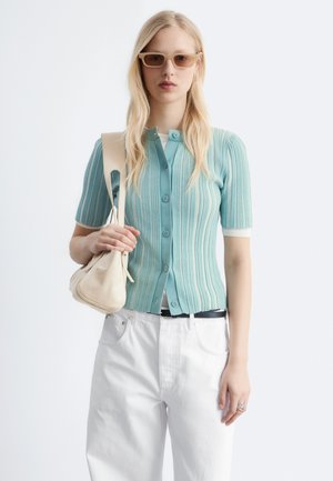 Mladá žena nosí rebrovaný tyrkysový krátky gombíkový kardigán, biele nohavice, béžové slnečné okuliare a nesie béžovú kabelku cez rameno.