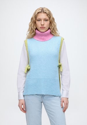 Un mannequin porte un col roulé rose, un gilet en maille bleu clair, une chemise blanche, un jean clair et des mocassins noirs, ainsi qu’un sac noir porté à l’épaule.