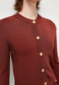 Cardigan a coste di colore ruggine con scollatura rotonda, dotato di bottoni in legno e design aderente. Tessuto testurizzato con dettagli di motivo sottili.