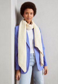 Jeune femme aux cheveux courts et bouclés, portant des boucles d'oreilles en créole, une écharpe blanche, un blazer lavande, un haut blanc et un jean bleu clair, debout contre un mur gris.