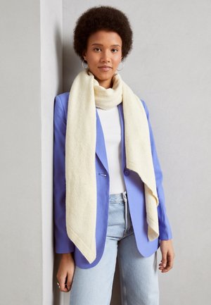 Jeune femme aux cheveux courts et bouclés, portant des boucles d'oreilles en créole, une écharpe blanche, un blazer lavande, un haut blanc et un jean bleu clair, debout contre un mur gris.