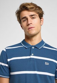 Polo shirt blu con righe orizzontali bianche, dotato di colletto e patta con due bottoni. Include un piccolo logo ricamato Lee sul petto.