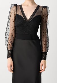 Body nero con scollatura a V profonda, dettagli in pizzo e maniche a sbuffo trasparenti con pois. I polsini in pizzo nero completano il design.