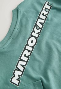 Mátově zelené tričko s kulatým výstřihem, se silným černým grafickým nápisem "MARIOKART" na levé straně. Hladká bavlněná textura s krátkými rukávy.