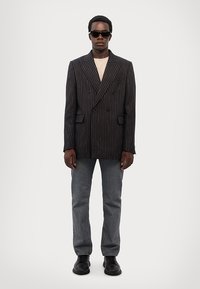 Dobbeltbrystet stribet blazer i sort og grå med to forlommer, båret over en beige top, parret med grå jeans og sorte sko.
