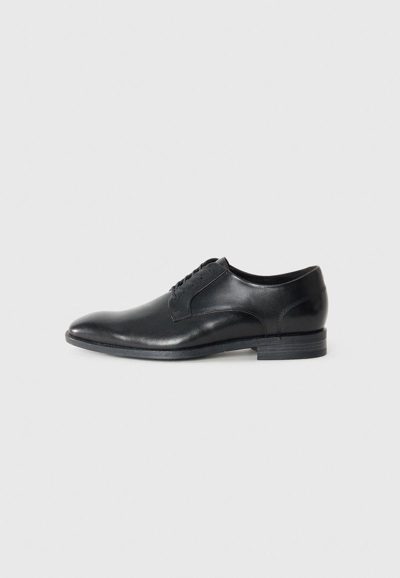 Black leather dress shoe with a rounded toe, minimalistic design, five eyelets for laces, and a flat rubber sole. Smooth texture.