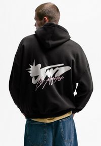 Črna kapa s kapuco z velikim srebrnim motivom in roza napisom na hrbtni strani. Mehka tkanina z udobnim ohlapnim krojem in žepom kenguru.