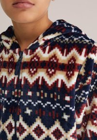 Fleece overal s kapucí s geometrickým vzorem v tmavě modré, červené a smetanové barvě, s plným zipem a měkkou texturou.