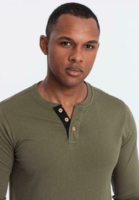 Man med kort hår som bär en långärmad henley-tröja i olivgrön med tre beige knappar och ett svart innerfoder.