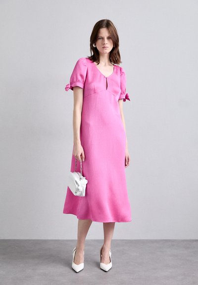 Puffärmad rosa klänning med en strukturerad yta, knä-lång fåll och en nyckelhålsdetalj fram. I kombination med vita högklackade skor och en liten vit väska.