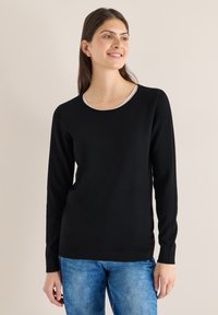 Femme souriante, portant un pull noir à manches longues et un jean bleu, debout devant un fond uni de couleur claire.